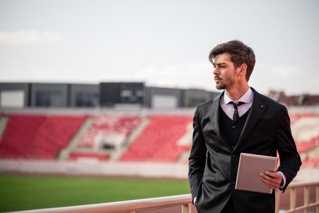 A man in a suit looks on to a football pitch from the stands