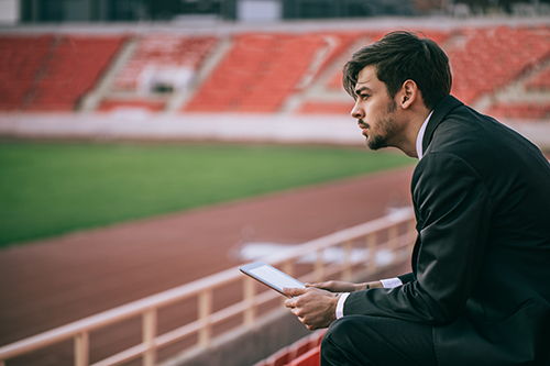 A man sits in the stands of a football pitch holding a tablet
