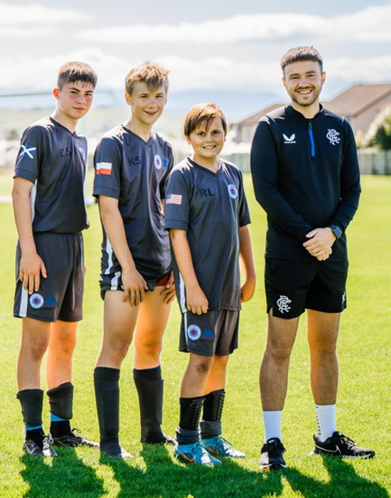 Three young footballers stand on a pitch with their coach