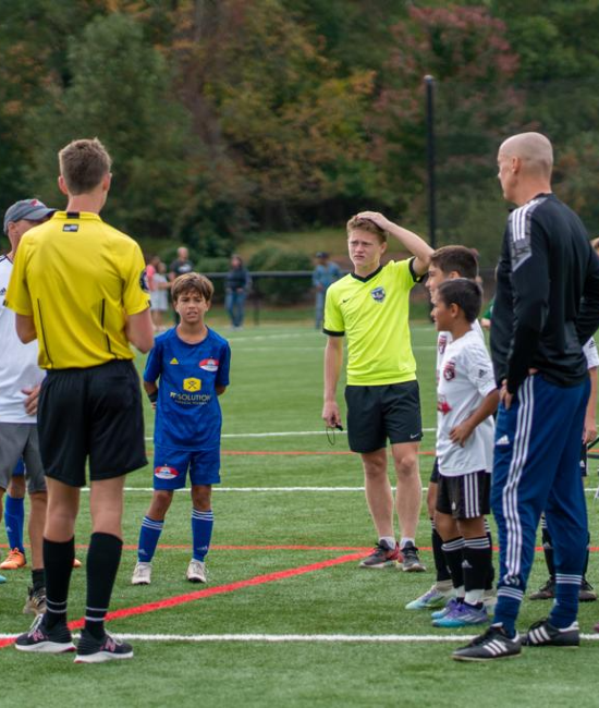Two football officials speak with a group of young footballers on a grass pitch